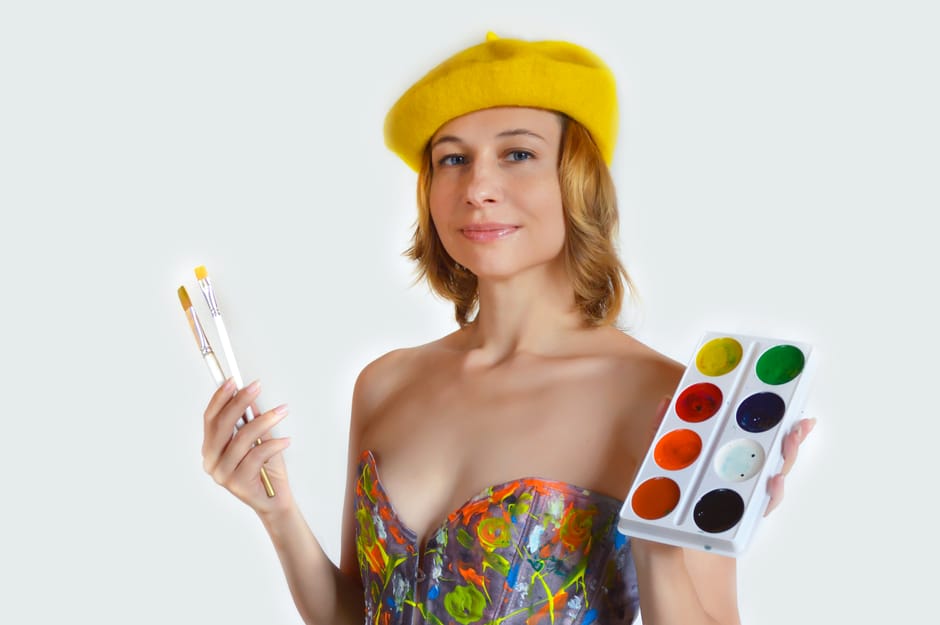 Delighted young female artist in colorful wear and yellow beret with brushes and palette of watercolor standing against white background and looking at camera