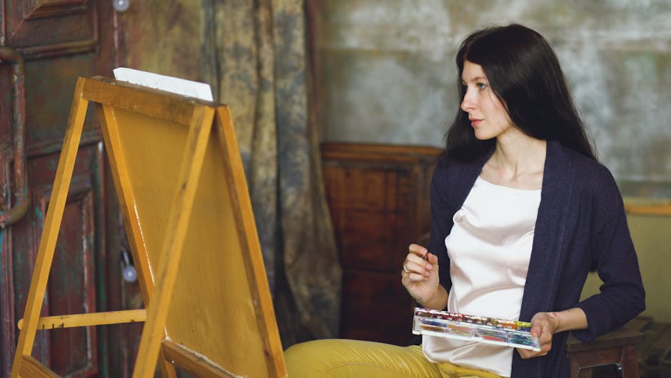A woman artist with long black hair paints on an easel indoors, holding a palette with focused expression.